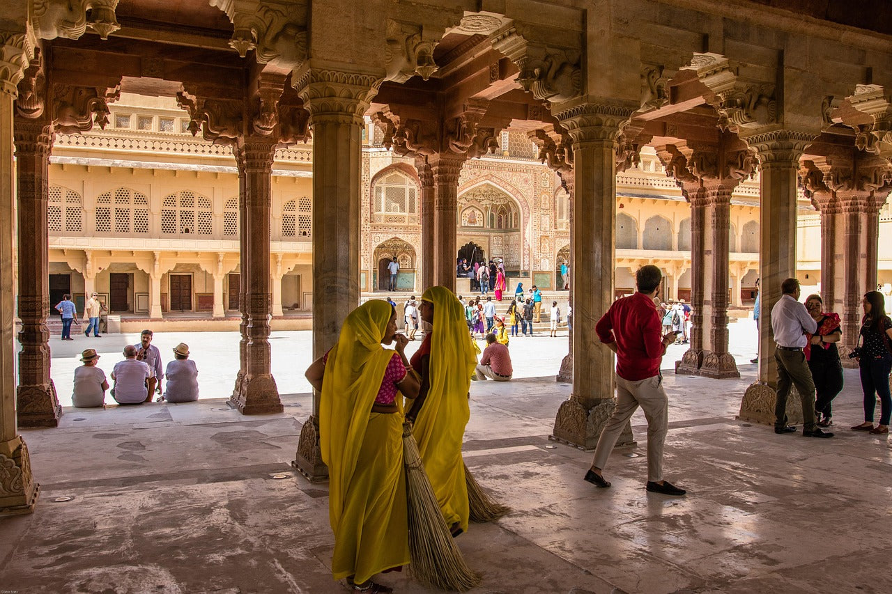 People in a courtyard with ornate architecture, including women in yellow saris.